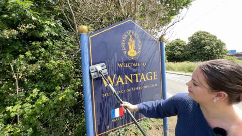 Ellie, who has brown hair tied-up, is using a mop to clean a large blue road sign welcoming motorists in to Wantage.