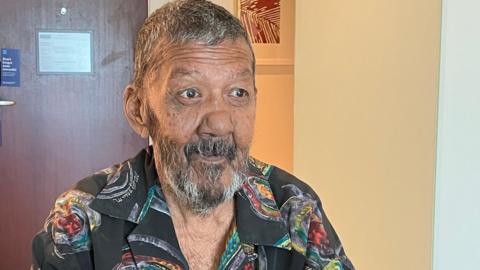 Nizam Hosein with short greying hair and bread looking off camera. He is wearing a black shirt with a mythical, colourful pattern