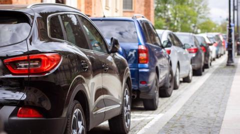 A line of cars are parked on a cobbled street. The cars can be seen going into the distance.