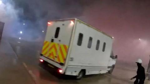 A screenshot of a police van seen on the Elbit Systems UK site. The van is a large white vehicle with small windows and a chevron pattern across the back. A figure wearing black with a white helmet can be seen in the lower right corner of the image holding a long unidentified object. 