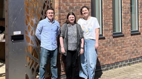 A man in a blue shirt, woman wearing grey and another woman in a white top stood outside a red brick building. They're all smiling.