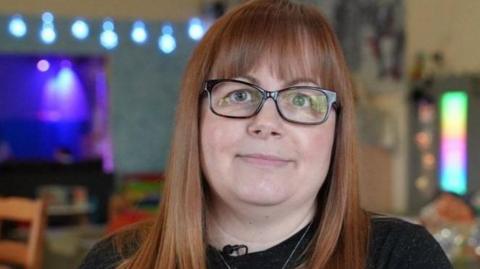 A head-and-shoulders picture of Lynn Hammel-Randall. She has long brown-red hair and a fringe. She is wearing a black jumper, glasses with a dark rim and a necklace with a cross pendant. She is sat in the cafe and the background is blurred.