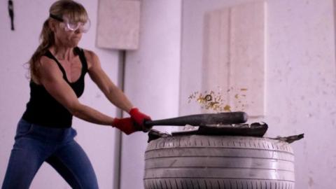 Picture of a woman in a rage room smashing tyres with baseball bats. She is wearing a visor, black vest top and jeans.