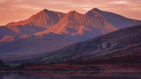 Yr Wyddfa seen from a distance, with a lake at the foot of the picture and sunshine on the upper part of the mountain