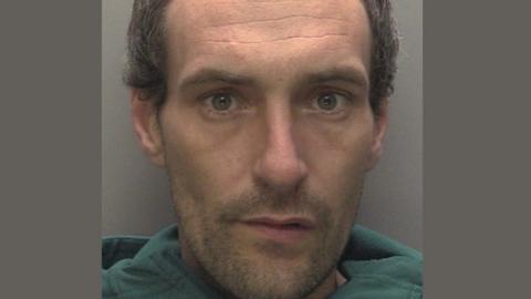 A police mugshot of Ashley Moss. He has dark wavy hair and a short beard and is wearing a green top, He is looking straight towards the camera