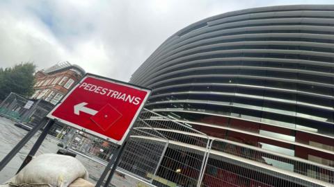 Exterior of the Curve theatre in Leicester with a panel missing from its sunbreaker and fencing in front of it, including a red sign directing pedestrians in front of the building