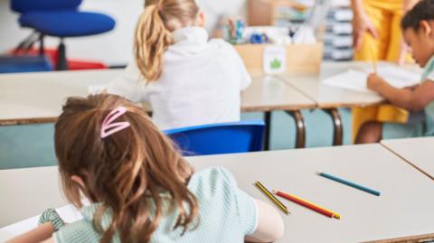 Three girls in primary school writing at their desks. They are all wearing green dresses and are pictured from the back. 