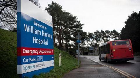 A sign outside the William Harvey Hospital points the way to the emergency departments, car parks and other sites. There is a bus on the road which has double yellow lines. The sign stands on a grass slope.