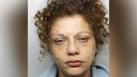 Katrina Dixon staring at the camera for her mugshot, with curly brown hair in a ponytail and scars on her face with puffy eyes. 