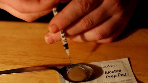 A man sits in front of a light brown wooden table, holding a hypodermic needle over a teaspoon containing a brown liquid. The spoon is on top of a white sealed packet with the words 'Alcohol Prep Pad' printed on it in blue lettering. 