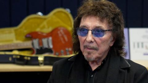 Man with blue-tinted glasses, beard and dark hair in a black jacket with black shirt. Behind him is a red guitar