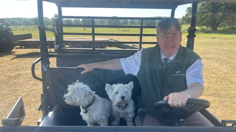 Colin Rayner is driving a quad bike with his two West Highland terrier dogs sitting to his right hand side as he holds the wheel. Grass fields can be seen behind them.