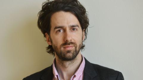 Jamie Bartlett - Radio 4 Image from 2022. He is wearing black suit jacket with pink shirt