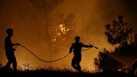 Vegetation is on fire and two people are holding a hose dowsing the flames 