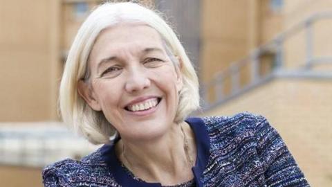 A woman with mid-length blonde hair and wearing a blue jumper, leans to her the right as she smiles.