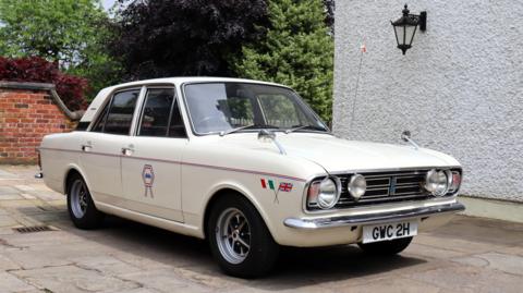 The white Ford Cortina parked next to a building, with the England football team emblem and British and Mexican flags on it