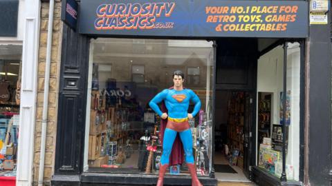 A full-sized, roughly 7-foot-tall statue of Superman stands outside on the high-street of a storefront of a retro toy shop named "Curiosity Classics”. The statue stands in a heroic, authoritative stance with hands planted firmly on his hips. He wears the classic suit with a cape. The statue stands on a grey, stone-textured rectangular block featuring the raised inscription "MAN OF STEEL."