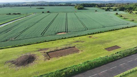 A strip of grass before an expanse of lush fields. There is a pile of earth and two rectangles dug out of the grass.