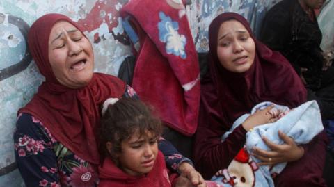 Two women cry out as they hold a child and a baby wrapped in a blanket, next to a wall in a school for displaced people in Gaza