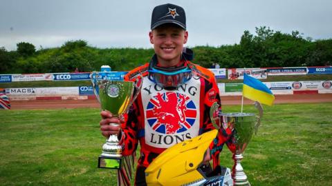 Cooper Rushen standing trackside with two silver trophies and his yellow helmet, wearing Young Lions jersey & black baseball cap
