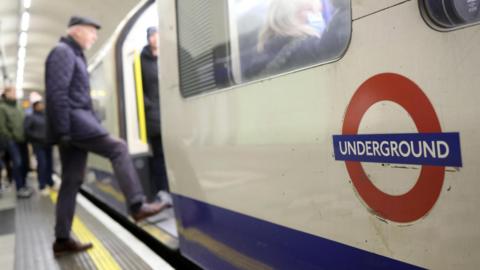 A close-up of a London Underground logo on the side of a train. In the background a man is stepping on to the train.