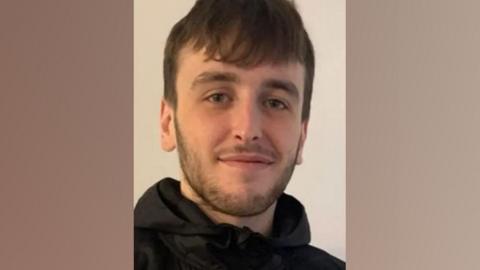 Sam Rimmer, who has short brown hair and short stubble, smiles at the camera while wearing a black coat