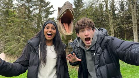 Shini and Joel in the foreground of a photo. They look terrified and are running away from a t-rex.