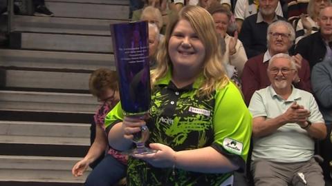 Katherine Rednall, wearing a bright green and black top, holds the World Indoor Bowls title trophy with spectators sitting in the background