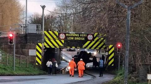 A bridge with yellow and black signs around it warning road users it is a 'very low bridge'. Underneath it a vehicle looks to be on its side. Two people in orange high visibility outfits are walking towards the blockage under the bridge. 