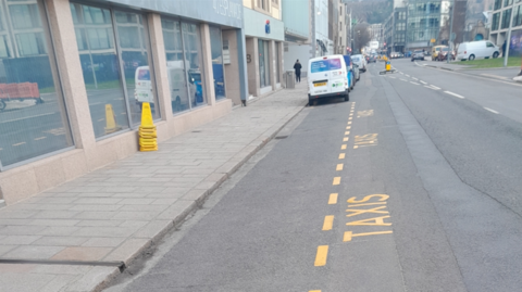 A new taxi rank on the esplanade - a long stretch of road with buildings to the left and right, heading into town. In the foreground is road and pavement, the taxi rank is marked by yellow dashes and the repeated word 'taxis'. Cars are parked further down the street. 