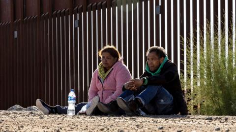 Two migrants sit on the ground near the border between the US and Mexico