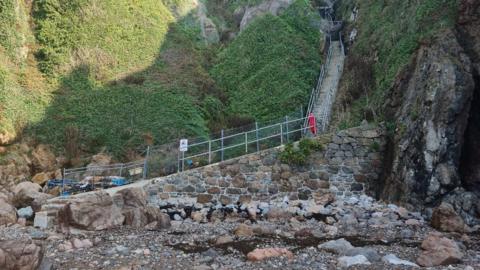 The bottom of the Petit Port steps leading on to a stony beach. There are metal handrails, fencing with a sign showing a hazard warning of falling rocks and a life buoy. The steps lead sharply up a stiff grassy cliff face. 
