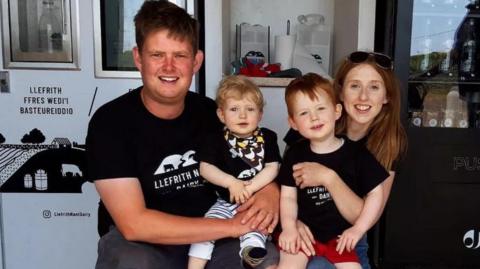 Llun o deulu, mam, dad a dau fab ifanc yn gwenu. Maen nhw'n gwisgo crysau-t du.
