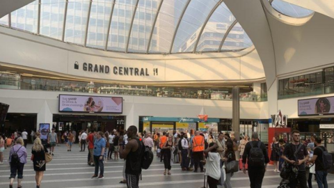 Approximately 30 people spread out across the station concourse on a typical day at New Street. Above them is a clear roof revealing the city-scape beyond. Some of the people face an arrivals and departures board which is out of shot.