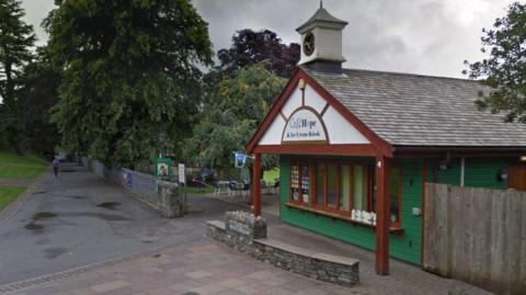 A green-and-white kiosk in a park with a clock tower and a sign saying Cafe Hope & Ice Cream Kiosk. Outdoor tables and chairs can be seen in the background along with trees and a path.