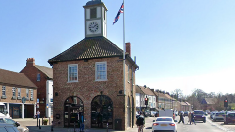 Yarm Town Hall in the sun. It is a square building with a flagpole attached to the side and a small clocktower. Arched windows are on the ground floor with white framed windows on the first floor.