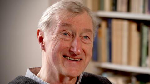 Julian Barnes smiling, top of checked shirt collar & dark top. Book shelf behind him.