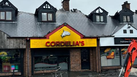 Cordelia's fish and chip shop - with a yellow and red sign - is badly damaged by fire. Above it is a slate roof with bay windows, badly damaged