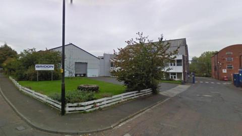 Two metal corrugated buildings stand on a corner street, there is a tree in front and to the left. A sign that reads 'Bridon' is positioned in grass on the left. 