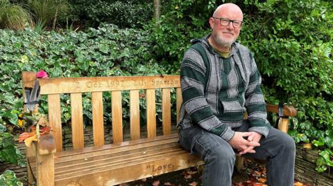 Simon Sparrow sits on a park bench, surrounded by trees and bushes. There are flowers and some feathers attached to the arm of the bench