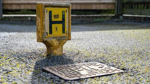 Close-up of a yellow fire hydrant marker post labelled ‘H 100 / 1’ next to a metal street drain on a paved surface.