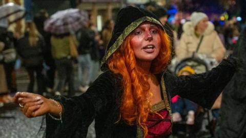 Shows a woman in red wig and black, pink and gold outfit dancing with people in the background holding umbrellas
