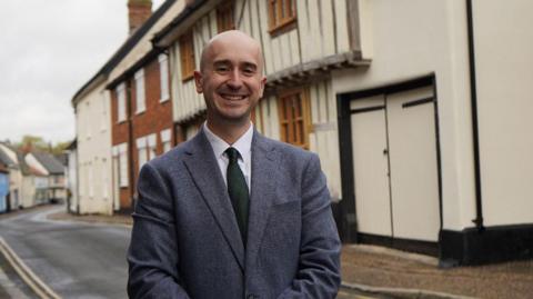 Ben Goldsborough is wearing a grey suit and standing outside a row of homes, one of which appears to be from the tudor-era.