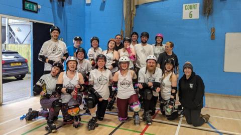 About 20 people in roller skates, knee pads and helmets pose together. Some are smiling and some are pulling silly faces.