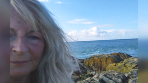 Di Mills looking into the camera on the island, with rocks and the sea behind her