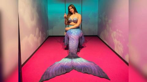 A woman sitting on a rope swing in a small room where the walls and floods are painted pink. She's wearing a shell bra and a large mermaid tail over her legs, in shades of iridescent blues, pinks, purples and blues.