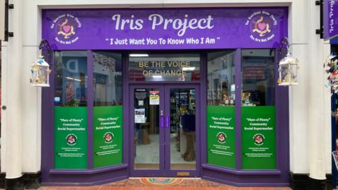 The front of a community social supermarket in Nuneaton. The  door and window frames are painted purple and the sign above the front door says 'Iris Project' with the message underneath saying 'I just want you to know who I am'.