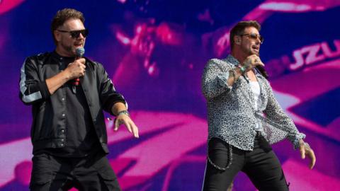 Two male singers stand on a large concert stage in front of a bright pink and purple LED screen. Both wear sunglasses and hold handheld microphones. One is dressed in a black jacket and black trousers, while the other wears a patterned shirt over a white top with black trousers and a chain at his waist. Stage lighting and part of a band setup are visible in the background.