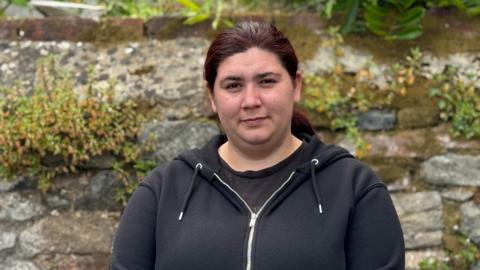 A woman in her mid-20s wearing a black hoodie and a black top. She is stood in front of a stone wall with plants growing out if it