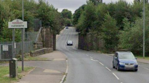 A road in Barnsley, with a couple of cars using the route. The road narrows in the distance due to brick walls on both sides. A sign on the left reads: "Cudworth."